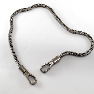 Könnte beinhalten: Eine silberne Kette mit zwei silbernen Verschlüssen. Die Kette ist in einem geflochtenen Design gefertigt.