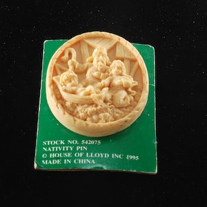 May include: A cream-colored ceramic nativity scene pin with a green background. The pin features a depiction of Mary, Joseph, and baby Jesus in a manger. The text on the card reads "Stock No. 542075 Nativity Pin © House of Lloyd Inc 1995 Made in China."