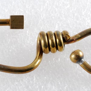 May include: A gold-colored metal hook with a coiled section in the middle. The hook has a square-shaped piece at one end and a rounded end at the other.
