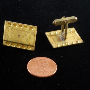 Puede incluir: Un par de gemelos de color dorado con un diseño rectangular. Los gemelos tienen una superficie texturizada y un borde elevado.