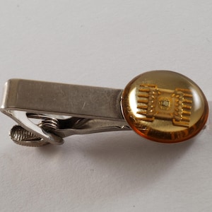 May include: A silver-toned tie clip with a gold-colored, circular accent. The accent features a detailed, gold-colored design, possibly a circuit board or similar technological element. The clip is designed to secure a tie.
