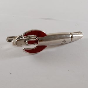 May include: Silver-toned rocket-shaped tie clip with red accents. The clip features a streamlined design with a pointed nose and red fins. A stylish accessory for a formal or casual outfit.