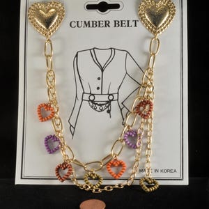 May include: Gold-tone metal cummer belt with heart-shaped clasps and dangling heart charms in orange, purple, and green. The card reads "CUMBER BELT" and features a line drawing of a dress with a belt. A penny is at the bottom.