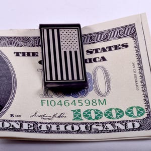 May include: A black metal money clip featuring a vertical American flag design in silver. The clip is holding a one thousand dollar bill, which displays the text "THE UNITED STATES OF AMERICA", "ONE THOUSAND", and the serial number "FI0464598M".