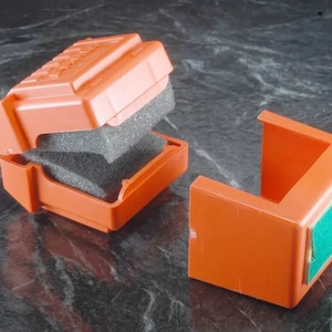 May include: Two orange plastic boxes with a foam insert. One box is open and the other is closed. The closed box has a green sticker on the side.