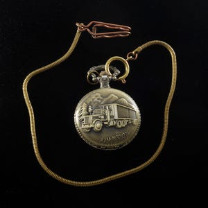 Può includere: Un orologio da tasca dorato con una catena. Il quadrante dell'orologio presenta un'immagine incisa di un camion che guida attraverso un passo di montagna.