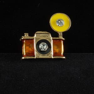 Peut inclure: Broche en forme d'appareil photo doré avec des accents orange et noirs. L'appareil photo est doté d'un flash jaune et de deux strass clairs et étincelants. Un accessoire d'inspiration vintage.