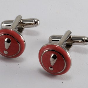 Puede incluir: Un par de gemelos de plata con un círculo de esmalte rojo que presenta un signo de exclamación blanco.