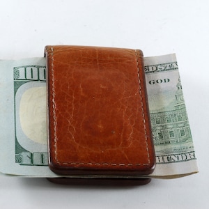 Könnte beinhalten: Ein brauner Leder-Geldclip, der zwei Hundert-Dollar-Scheine hält. Der Geldclip ist rechteckig mit einer strukturierten Oberfläche und einem Magnetverschluss. Die Geldscheine sind auf beiden Seiten des Clips sichtbar.