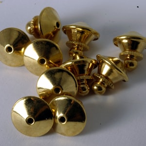 May include: A group of ten gold-toned metal pin backs. The pin backs are round with a small hole in the center. They have a raised, domed top and a small, round pin on the bottom.