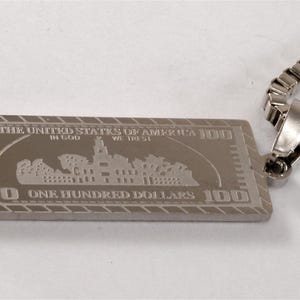 May include: A silver-colored rectangular pendant with the design of a $100 bill, attached to a silver chain. The pendant features the text "THE UNITED STATES OF AMERICA" and "ONE HUNDRED DOLLARS".