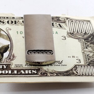 May include: A silver money clip with a braided design on a $10,000 bill. The bill has the text "THE FACE OF THE UNITED STATES OF AMERICA" and "SERIES 1990" printed on it.