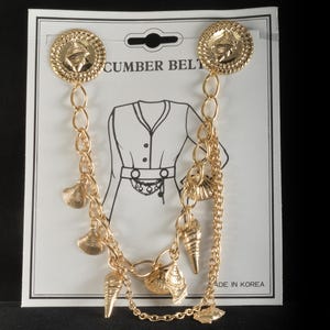 May include: Gold-toned cummerbund belt with seashell charms. The belt features two large round decorative clasps and a chain with various seashell pendants. The card reads "CUMBER BELT" and "MADE IN KOREA".