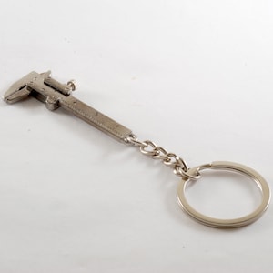 Könnte beinhalten: Ein silberfarbener Messschieber-Schlüsselanhänger. Das Miniaturwerkzeug hat ein detailliertes Design mit einer Kette und einem Schlüsselring. Der Messschieber hat ein Lineal entlang seiner Länge. Ein praktisches und stilvolles Accessoire.