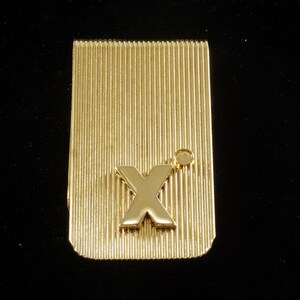 Puede incluir: Pinza para billetes dorada con una letra 'X' en relieve en el centro. La pinza tiene una superficie estriada.