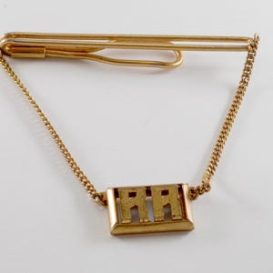 May include: A gold tie clip with a chain and a rectangular pendant with the letters "AA" engraved on it.