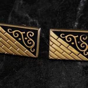 May include: A pair of gold cufflinks with a black enamel design. The design features a geometric pattern and a stylized floral motif.