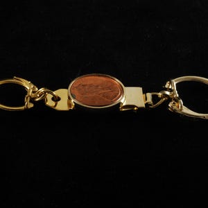 May include: A gold-colored metal keychain with a copper-colored coin in the center. The coin is oval-shaped and has a detailed design. The keychain has two key rings, one on each end, and a rectangular bar in the middle.