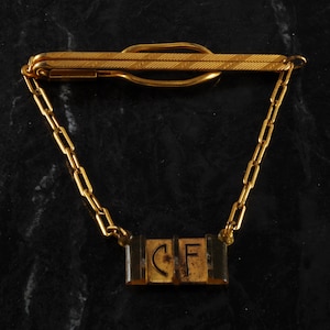 May include: A gold tie bar with a chain and a rectangular tag with the letters "CF" engraved on it.