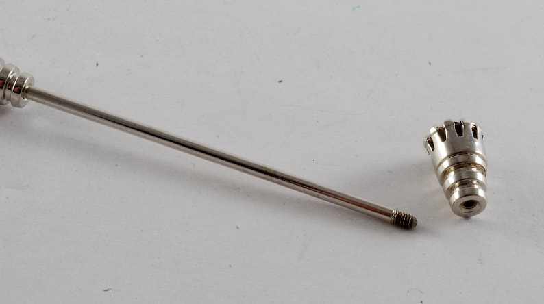 May include: A silver metal rod with a threaded end and a decorative, threaded, crown-shaped tip. The rod and tip are separated.