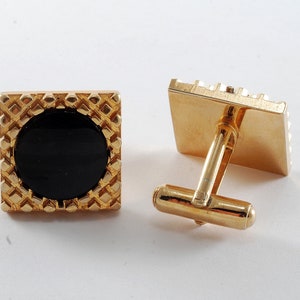 Può includere: Un paio di gemelli d'oro con un design quadrato nero. I gemelli hanno una superficie testurizzata e una forma quadrata.