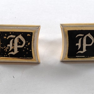 May include: A pair of gold cufflinks with a black enamel background and a white letter "P" in a gothic font.