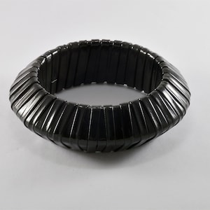 Könnte beinhalten: Ein schwarzes, dehnbares Armband mit geripptem Design. Das Armband besteht aus rechteckigen, dunkel gefärbten Segmenten, die eine strukturierte, kreisförmige Form bilden. Das Armband wird auf weißem Hintergrund gezeigt.