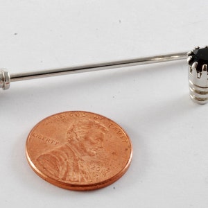 May include: A silver tie tack with a black gemstone set in a crown-like design. The tie tack is about the size of a US penny.