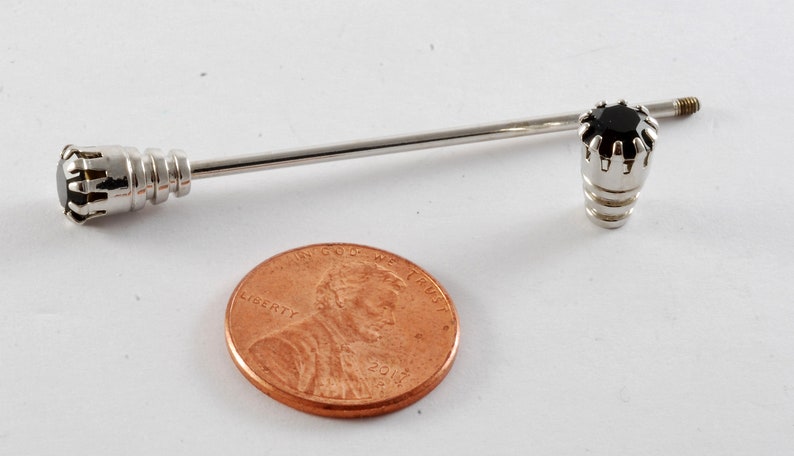 May include: A silver tie tack with a black gemstone set in a crown-like design. The tie tack is about the size of a US penny.