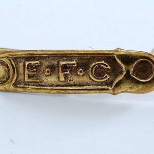 Könnte beinhalten: Eine goldfarbene Metallnadel mit den eingravierten Buchstaben "E.F.C.".