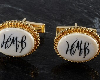 May include: A pair of gold cufflinks with a white oval inlay. The inlay has the initials "H.M.B." written in black ink.