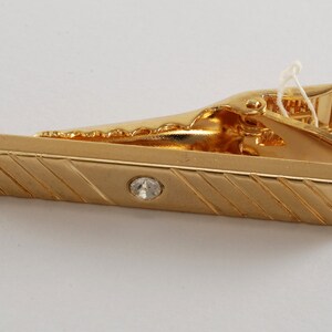 Könnte beinhalten: Goldene Krawattenklammer mit einem klaren Strassstein in der Mitte und einem gestreiften Design. Die Klammer ist geschlossen.