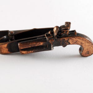 Könnte beinhalten: Eine Krawattenklammer im Vintage-Stil in Form einer Pistole. Die Krawattenklammer ist aus bronzefarbenem Metall gefertigt und zeigt detaillierte Elemente einer Steinschlosspistole. Die Klammer ist ca. 5 cm lang und hat einen Federverschluss.