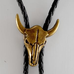 May include: A black bolo tie with a gold-toned bull skull pendant. The pendant has a detailed design and a textured finish.