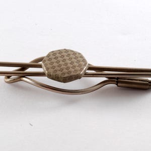 Könnte beinhalten: Eine Vintage-Krawattenklammer mit einem geometrischen Muster auf dem zentralen sechseckigen Teil. Die Klammer besteht aus einem gedämpften, metallischen Material und hat ein schlichtes, funktionales Design. Der Hintergrund ist schlicht weiß.