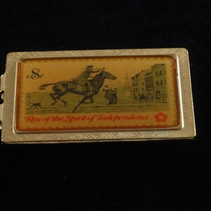 Könnte beinhalten: Ein rechteckiger, goldfarbener Geldclip aus Metall mit einem rechteckigen Bild eines Reiters auf einem Pferd, eines Hundes und von Gebäuden. Das Bild trägt die Aufschrift "Rise of the Spirit of Independence" und "8¢".