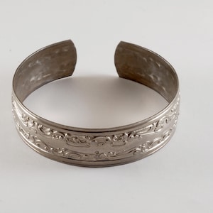Könnte beinhalten: Silberfarbenes Armreif-Armband mit offenem Design. Das Armband hat ein verziertes, geprägtes Blumenmuster um das Band. Das Metall hat ein leicht gealtertes Aussehen, was seinen Vintage-Charme unterstreicht. Ein klassisches Schmuckstück.