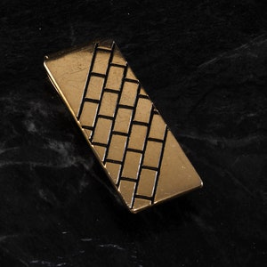 May include: A gold-colored metal money clip with a black brick pattern design.