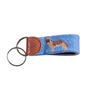 May include: Blue German Shepherd dog keychain with a brown leather accent and silver key rings. The dog is embroidered on a blue background. The leather accent has a logo. A practical and stylish accessory for dog lovers.