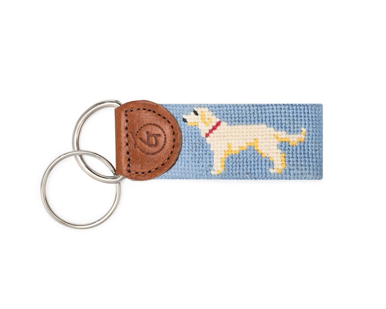 May include: A blue and white needlepoint key fob featuring a yellow Labrador Retriever design. The key fob has a brown leather strap with a metal key ring.