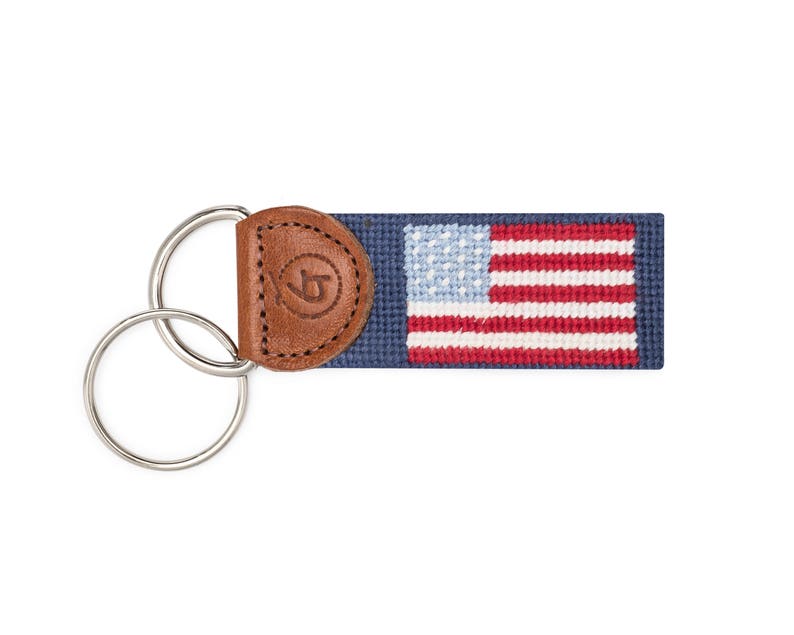 May include: A blue and white American flag keyring with a brown leather loop. The keyring has a silver double ring.