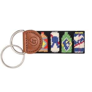 May include: A black needlepoint keychain featuring four colorful soda can designs. The keychain has a brown leather backing with a silver key ring. The leather backing has a stitched logo.