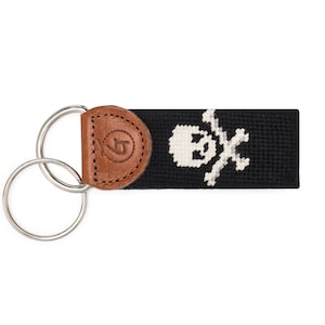 May include: A black and white skull and crossbones design on a black key fob with a brown leather accent. The key fob has a silver double ring.