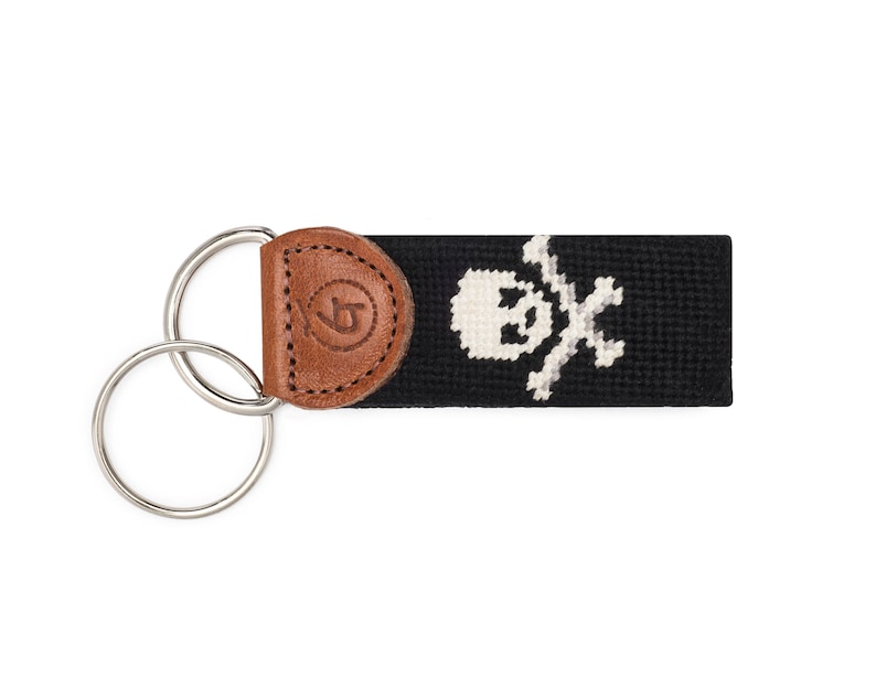 May include: A black and white skull and crossbones design on a black key fob with a brown leather accent. The key fob has a silver double ring.