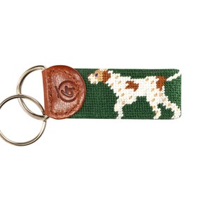 May include: A green and brown needlepoint key chain with a white dog design. The key chain has a brown leather loop with a logo.