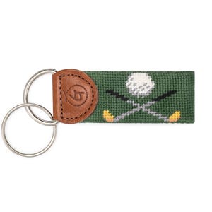 May include: A green and brown key chain with a golf ball and crossed golf clubs design. The key chain has a split ring and a leather fob with a logo.