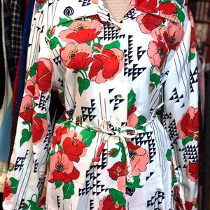 May include: A white long-sleeved shirt with a geometric pattern and red, pink, and green floral print. The shirt has a button-up front and a tie belt at the waist.