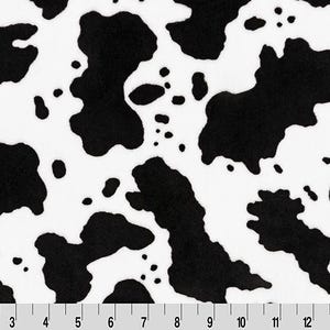 May include: A fabric featuring a black and white cow print pattern with irregular spots. A ruler is visible at the bottom edge, showing measurements in inches from 1 to 15, indicating the scale of the repeating animal print design.
