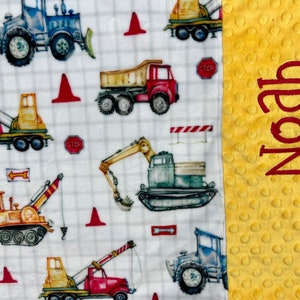 May include: A white and yellow baby blanket with a construction theme. The blanket features a variety of construction vehicles, including trucks, excavators, and cranes. The blanket also has a red stop sign and red traffic cones. The blanket is personalized with the name "Noah" on the yellow side.