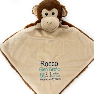 May include: A brown monkey plush toy with a tan blanket. The blanket has embroidered text: "Rocco Glen Grote 8 pounds 7 oz 21 inches 4:42 AM November 2, 2023". The monkey has a smiling face.
